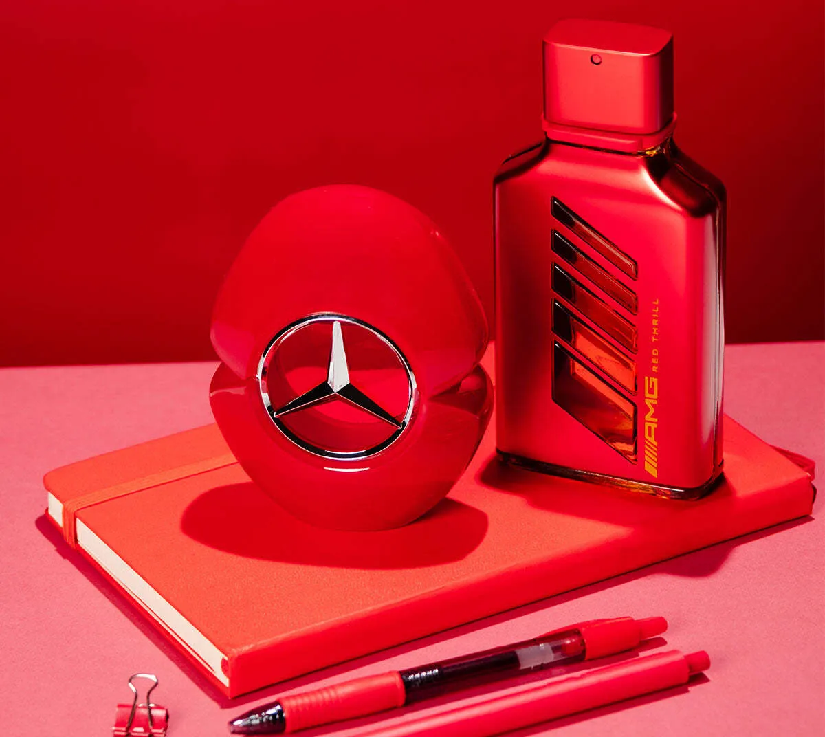 Mercedes-Benz perfume bottles, including AMG Red Thrill, displayed on a red notebook with matching red pens against a vibrant red background.